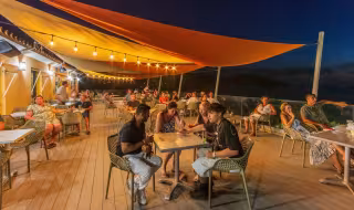 Terrasse du restaurant animée en soirée, sous les voiles d'ombrage et les guirlandes lumineuses