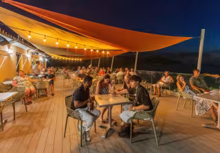 Terrasse du restaurant animée en soirée, sous les voiles d'ombrage et les guirlandes lumineuses