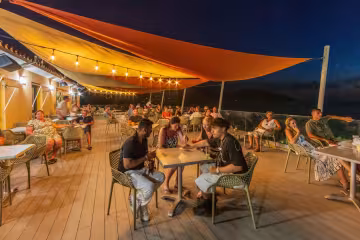 Terrasse du restaurant animée en soirée, sous les voiles d'ombrage et les guirlandes lumineuses